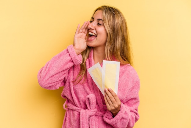 Young caucasian blonde woman wearing bathrobe holding depilatory bands isolated on yellow background  shouting and holding palm near opened mouth.