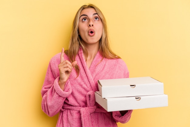 Young caucasian blonde woman wearing bathrobe holding pizzas isolated on yellow background  pointing upside with opened mouth.