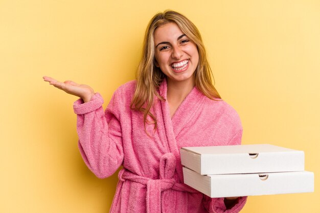 Giovane donna bionda caucasica che indossa accappatoio tenendo le pizze isolate su sfondo giallo che mostra uno spazio di copia su un palmo e tenendo un'altra mano sulla vita.