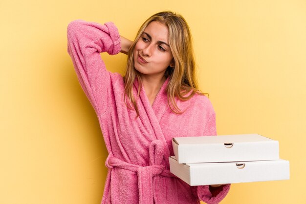 Young caucasian blonde woman wearing bathrobe holding pizzas isolated on yellow background  touching back of head, thinking and making a choice.