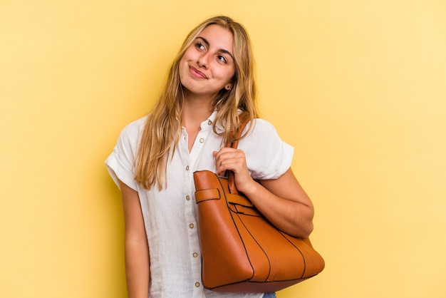 Young caucasian blonde woman wearing a leather bag isolated on yellow background  dreaming of achieving goals and purposes