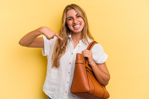 Young caucasian blonde woman wearing a leather bag isolated on\
yellow background person pointing by hand to a shirt copy space,\
proud and confident