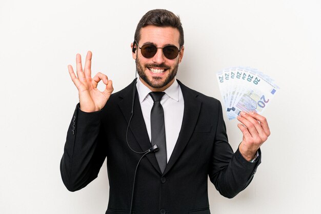 Young caucasian bodyguard man holding banknotes isolated on white background cheerful and confident showing ok gesture