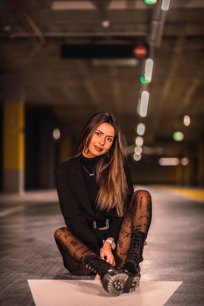 Young caucasian brunette in black jacket sitting on the ground in an empty underground car park. Night urban session in the city