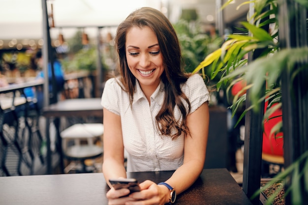 Young Caucasian brunette with big toothy smile sitting in coffee shop and using smart phone for reading or writing message.