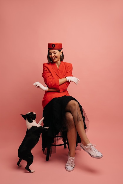 Young caucasian brunette woman in red outfit teaches her dog voice command while sitting on pink background Lifestyle and friendship concept