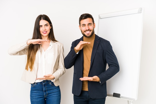 Young caucasian business couple isolated holding something with both hands, product presentation.