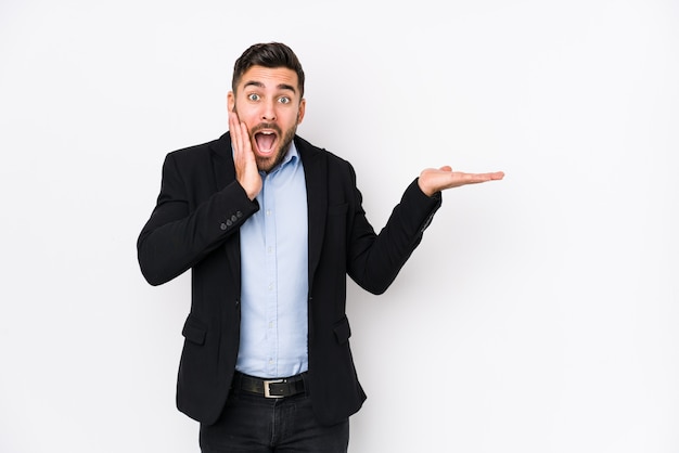 Young caucasian business man against a white wall isolated holds copy space on a palm, keep hand over cheek. Amazed and delighted.