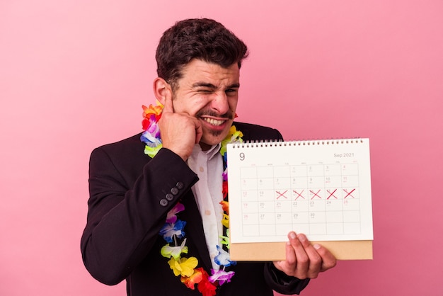 Young caucasian business man counting the days for vacations isolated on pink background covering ears with hands.