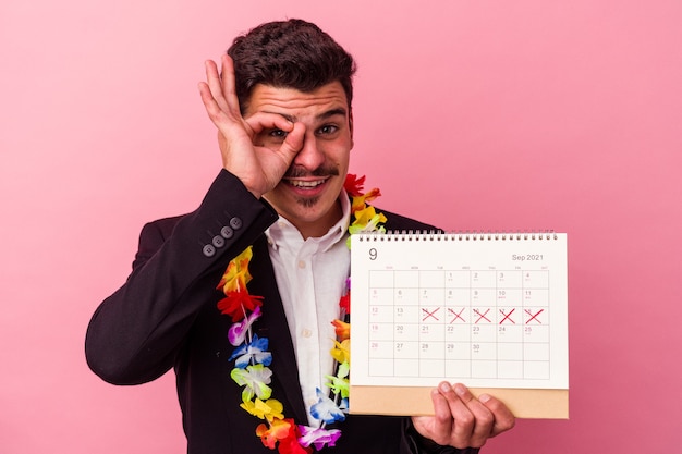 Il giovane uomo d'affari caucasico che conta i giorni per le vacanze isolato su sfondo rosa eccitato mantenendo il gesto ok sull'occhio.