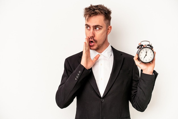 Young caucasian business man holding alarm clock isolated on white background is saying a secret hot braking news and looking aside