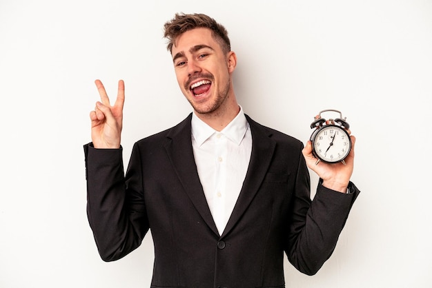 Young caucasian business man holding alarm clock isolated on white background joyful and carefree showing a peace symbol with fingers.