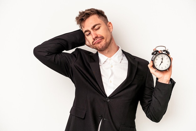 Young caucasian business man holding alarm clock isolated on white background touching back of head, thinking and making a choice.