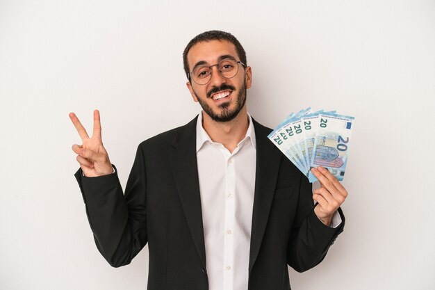 Young caucasian business man holding banknotes isolated on white background joyful and carefree showing a peace symbol with fingers.