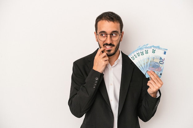 Young caucasian business man holding banknotes isolated on white background relaxed thinking about something looking at a copy space.