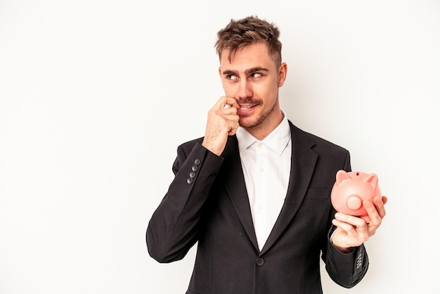 Young caucasian business man holding piggy bank isolated on white background relaxed thinking about something looking at a copy space.