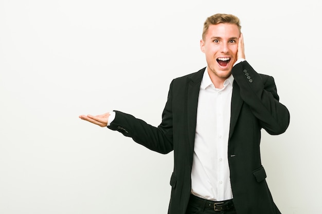 Young caucasian business man holds copy space on a palm, keep hand over cheek Amazed and delighted