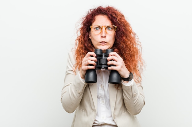 Young caucasian business redhead woman holding a binoculars biting fingernails, nervous and very anxious.