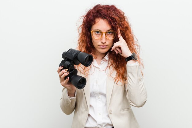 Young caucasian business redhead woman holding a binoculars pointing his temple with finger, thinking, focused on a task.