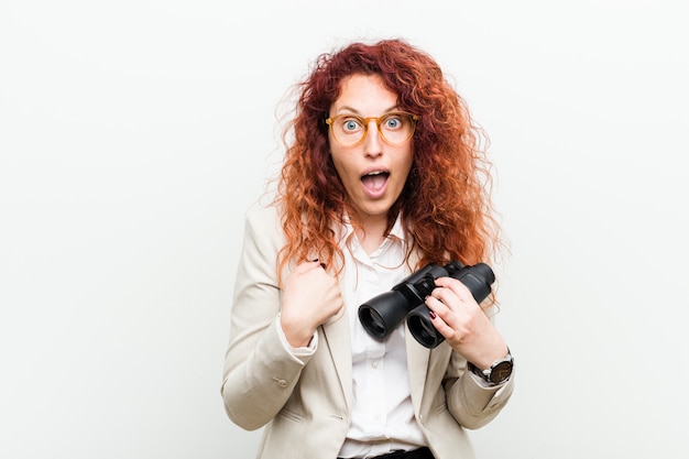 La giovane donna caucasica di affari di redhead che tiene un binocolo ha sorpreso indicare a se stessa