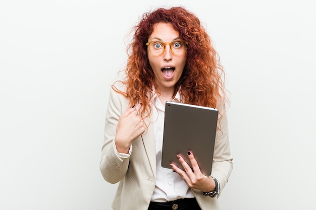 Young caucasian business redhead woman holding a tablet surprised pointing at himself, smiling broadly.