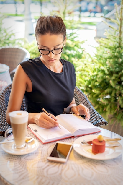 Young caucasian business woman in black dress writes in notebook sitting at table in cafe