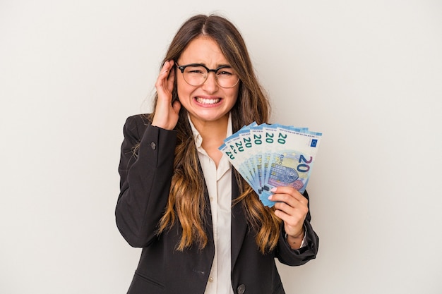 Young caucasian business woman holding banknotes isolated on white background covering ears with hands.