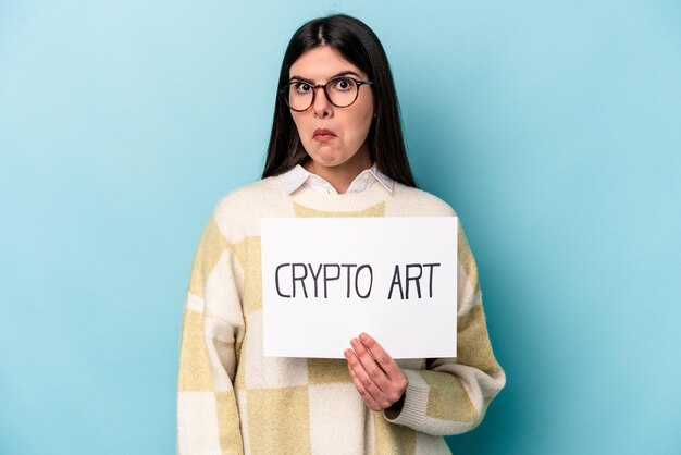 Young caucasian business woman holding a crypto art placard isolated on blue background shrugs shoulders and open eyes confused