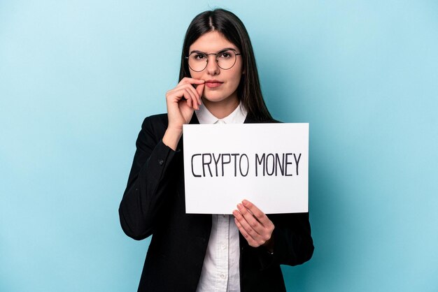 Young caucasian business woman holding a crypto money placard isolated on blue background with fingers on lips keeping a secret