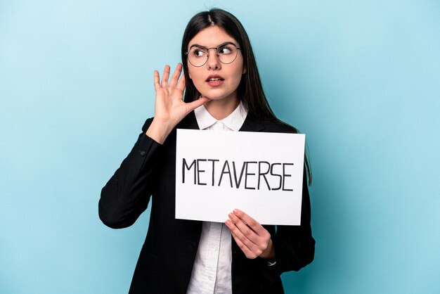 Young caucasian business woman holding a metaverse placard isolated on blue background trying to listening a gossip