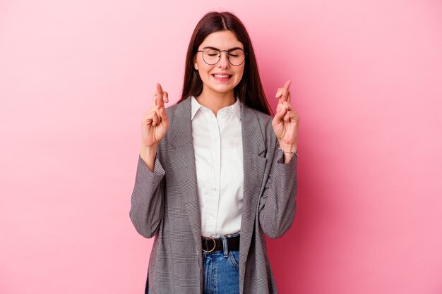 Young caucasian business woman isolated on pink background crossing fingers for having luck