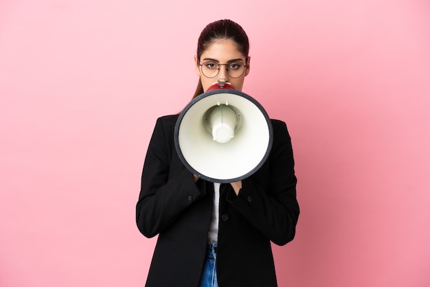 Young caucasian business woman isolated on pink background shouting through a megaphone