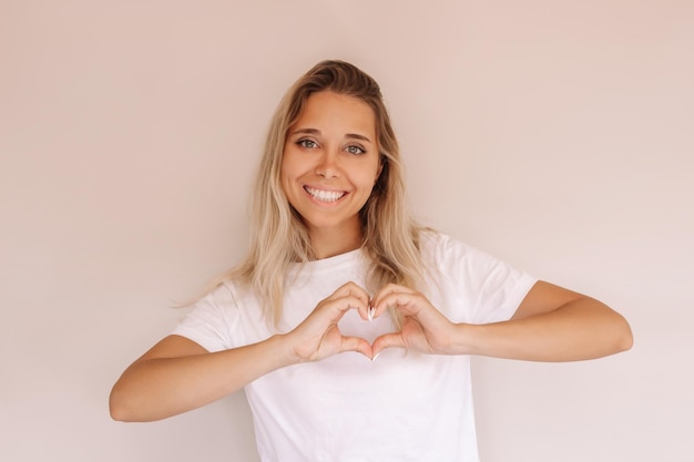 Young caucasian charming smiling blonde woman in white t-shirt forming a heart shape with her hands