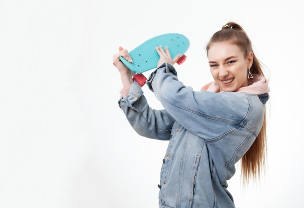 Young caucasian cheerful hipster woman in denim clothes with penny board on white background