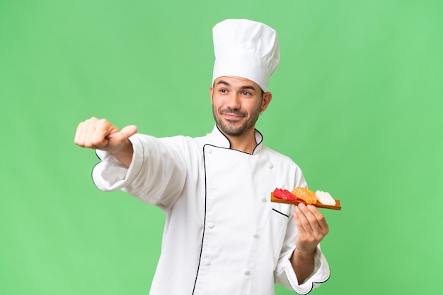 Giovane chef caucasico in possesso di un sushi su sfondo isolato dando un pollice in alto gesto