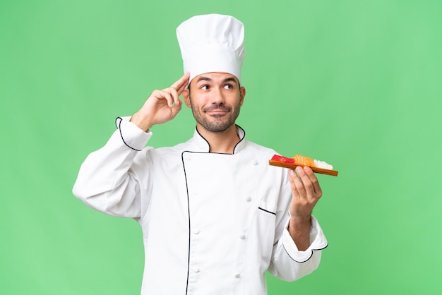 Young caucasian chef holding a sushi over isolated background having doubts and thinking