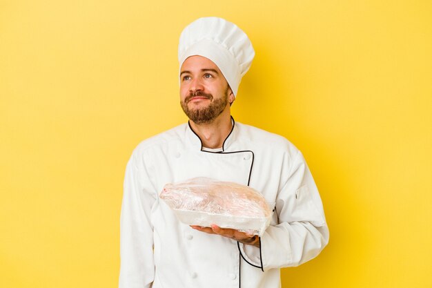 Young caucasian chef man holding chicken isolated on yellow background dreaming of achieving goals and purposes