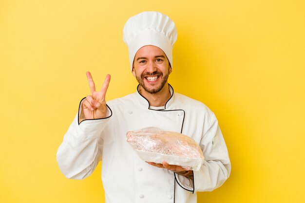 Young caucasian chef man holding chicken isolated on yellow wall showing number two with fingers.