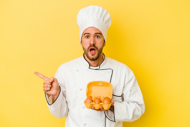 Young caucasian chef man holding eggs isolated on yellow background pointing to the side