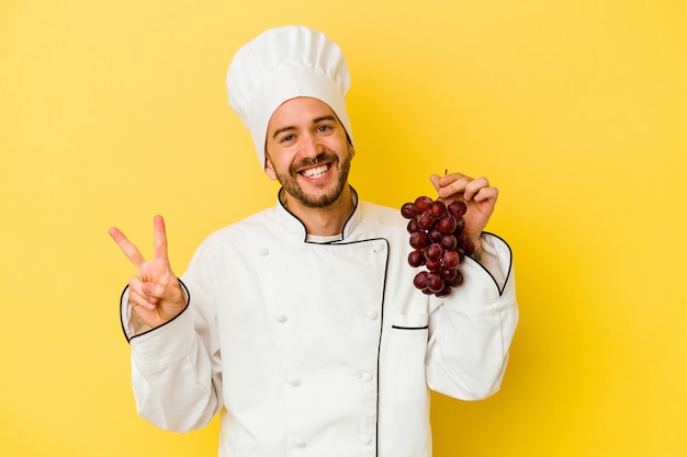 Young caucasian chef man holding grapes isolated on yellow background joyful and carefree showing a peace symbol with fingers.