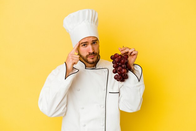 Young caucasian chef man holding grapes isolated on yellow wall pointing temple with finger, thinking, focused on a task.