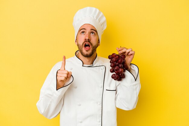 Young caucasian chef man holding grapes isolated on yellow wall pointing upside with opened mouth.