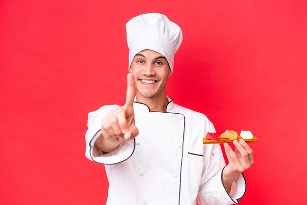 Молодой кавказский повар мужчина держит сашими на красном фоне, показывая и поднимая палец