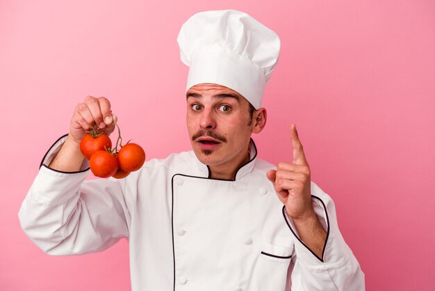 Young caucasian chef man holding tomatoes isolated on pink background having an idea, inspiration concept.