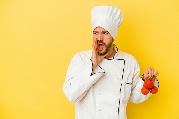 Photo young caucasian chef man holding tomatoes isolated on yellow background is saying a secret hot braking news and looking aside