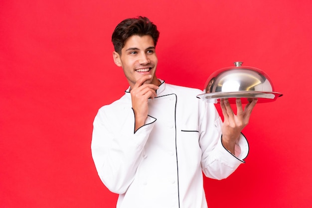 Young caucasian chef man holding tray isolated on red background looking up while smiling