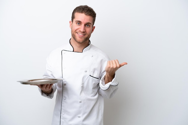 Young caucasian chef with tray isolated on white background pointing to the side to present a product