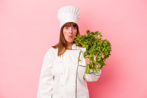 Young caucasian chef woman holding parsley isolated on pink background shrugs shoulders and open eyes confused.