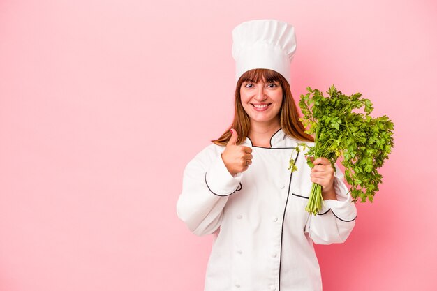 Young caucasian chef woman holding parsley isolated on pink background smiling and raising thumb up