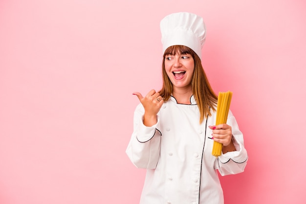 La giovane donna caucasica dello chef che tiene la pasta isolata su sfondo rosa punta con il pollice lontano, ridendo e spensierato.